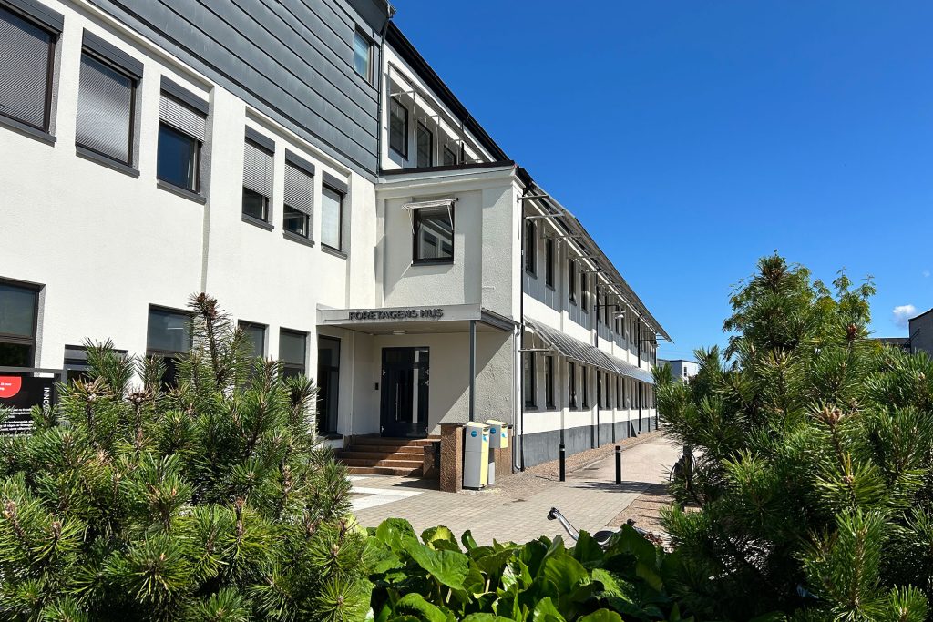 Vit byggnad med grönska och blå himmel. På bilden syns högra entrén.