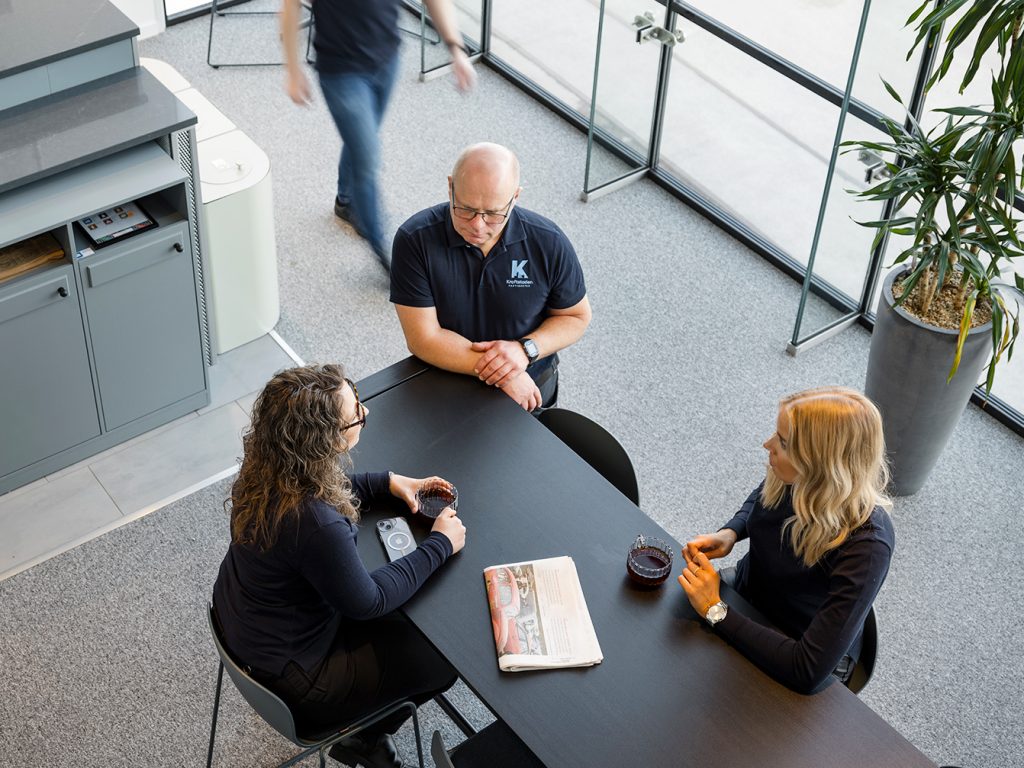 Tre kollegor diskuterar i luftigt kök med stora glasfönster. En person går förbi bakom.