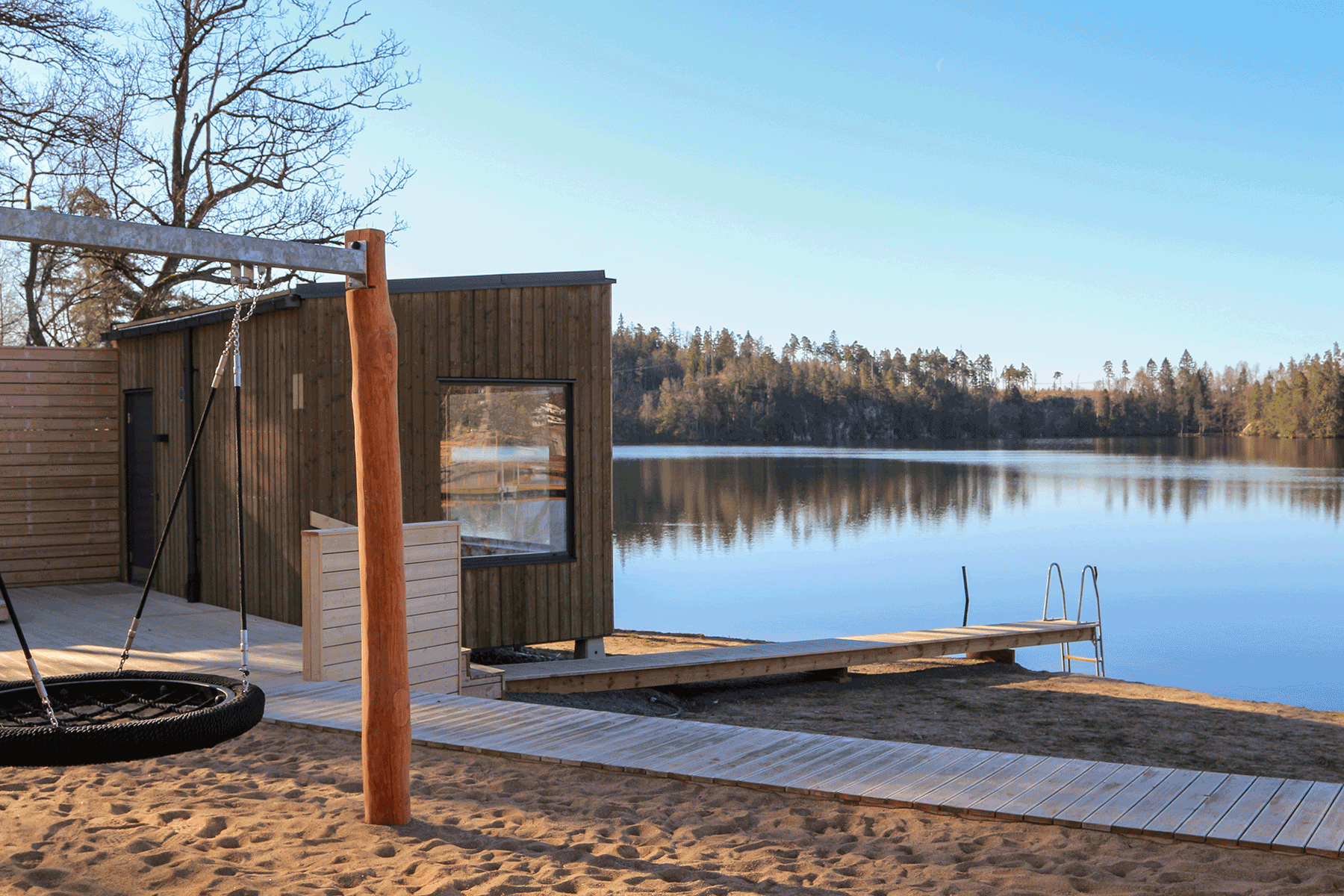 En lugn badplats vid en stilla sjö. I förgrunden syns en sandstrand med en rund gunga. En träspång leder till en modern träbyggnad med stora fönster och vidare ut till sjön. I bakgrunden syns en skog längs sjöns strand.