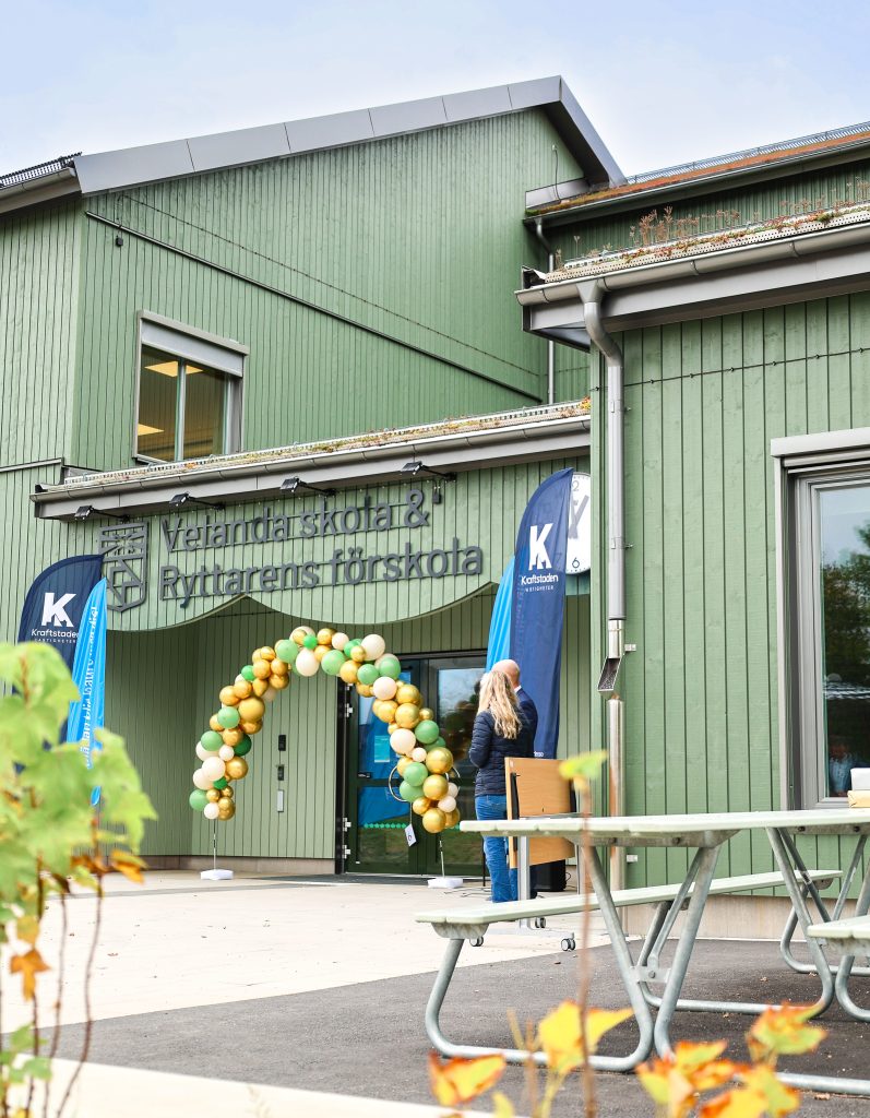 En skola med grön träfasad. Framför huvudentrén står en ballongbåge med ballonger i grönt, vitt och guld.